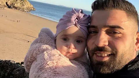 Alex Keleman Alex Keleman with baby Sophia pictured with the beach in Tenby in the background. He has short dark hair and a shirt beard and she is wearing a fluffy pink jacket and pink hat