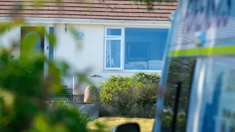 PA Media A detached bungalow with cream walls and a tiled roof is viewed from behind trees and shrubs. A large front window looks into a living room, while part of a police vehicle is visible at the edge of the image.