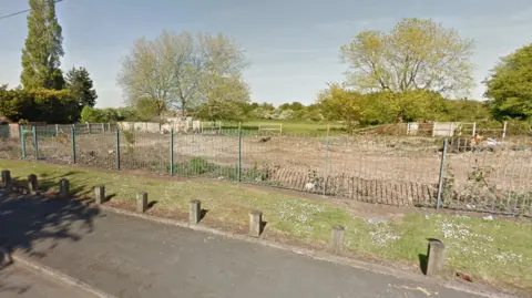Google An area behind green fencing, scraped back to the earth, with trees in the distance