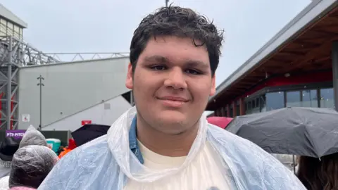 Will Khanna wears a plastic poncho in the Liverpool rain