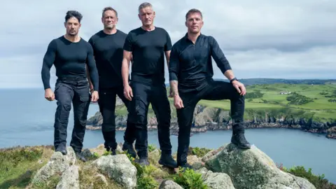 Pete Dadds | Channel 4 Four men in their black fatigues standing on a hilltop overlooking a coastline with a headland in the background and the sea beyond. The man on the right of the row perches his foot on a large rock. 
