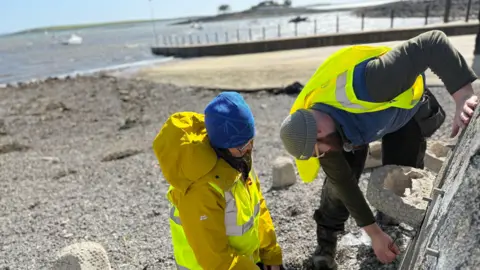 Mourne Gullion and Strangford officers Anna and Grant fitting a grey rock concrete rock pool. Anna is wearing a blue hat, yellow mustard coat and a yellow high vis vest with Mourne Gullion and Strangford logo. Grant is beside her wearing a blue puffy gilet with sunglasses hanging on the zip. He is also wearing a high vis jacket but it is not fully covering him. He has a green jumper under his puffer. The lough is in the baground. They are looking at the wall. 