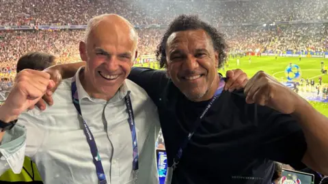 Tony Jones wearing a light blue shirt while holding his arm round the shoulders of co-commentator Don Goodman. They both have a fist in the air and are smiling. They are in the stands at the UEFA Champions League final, having just commentated on the match.