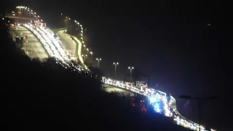 A view over a dual carriageway at night. One side of the road has cars lined up as far as you can see.