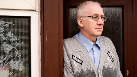 PA Media Billy Blue, a man with thinning white hair, glasses and a grey patterned top, with a blue shirt underneath it. He is standing in the doorway of his home, looking into the distance.