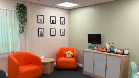 A view of a family room featuring two orange one-seater chairs with a round wooden table in between. To the right is a unit featuring three cupboards with a TV and coffee station on top.