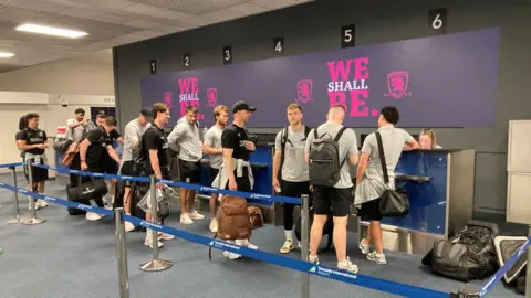 A dozen Middlesbrough players and team members stand at the check-in desks at the airport. The desks are numbered one to six, and a display underneath has the club's crest and the words: "We shall be." The team are wearing a mix of grey and black uniforms.
