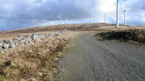 A gravel road, bordered by a stone wall on the left and a mound of earth on the right, winds its way through a fairly barren landscape to some wind turbines