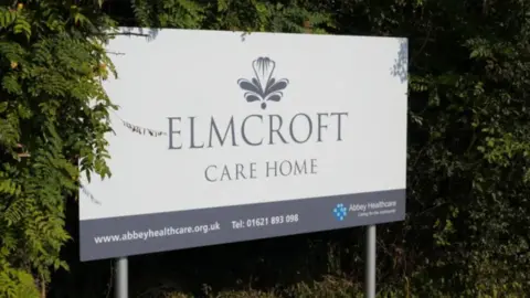 John Fairhall/BBC A sign that reads Elmcroft Care Home, installed in front of bushes or trees.