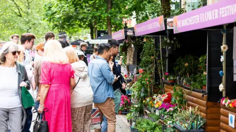 Carl Sukonik Dozens of people walk past or look at flower displays at a row of stalls. The stalls have Manchester Flower Festival signs above them.