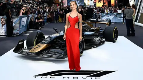 Getty Images Callie Cooke attends the European Premiere of F1: The Movie at Cineworld, Leicester Square. She is stood on white ground which has the movie's logo on it. She is wearing a red dress and a F1 car is behind her. There are several photographers and reporters in the background.