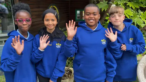 A group of four pupils, all holding a hand up and waving. They are all smiling and looking at the camera. They are all wearing blue hooded jumpers. They are outside by plants. 