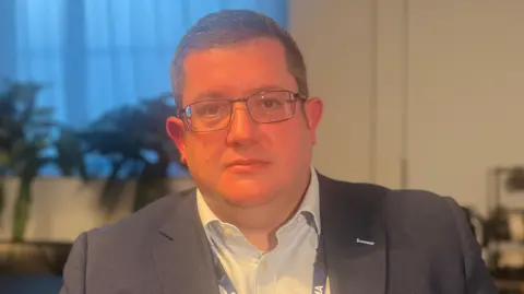 A man with dark hair and glasses, he is wearing a navy blazer and white shirt