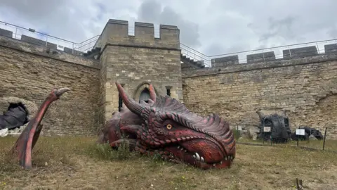 Eleanor Maslin/BBC Two large scaly dragon sculptures look as if they are bursting out of a castle's walls. On the left is the side-profile of a red dragon with a yellow eye and teeth protruding out of its mouth, as well as part of its wing with the rest submerged in the grass. Its tail appears to protrude out of the castle wall. On the right is a grey dragon, with a face, teeth and horns and big claws on its side.