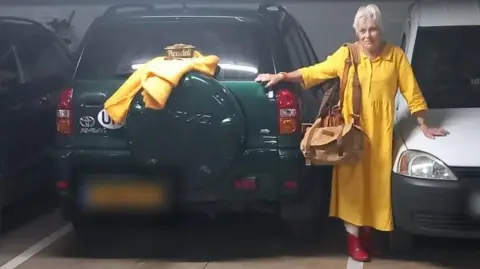 Aid Ukraine The image shows an indoor parking garage a green SUV with a yellow item draped over its spare tyre. A person wearing a yellow coat and red shoes is standing between the green SUV and a white van, holding onto both vehicles. 