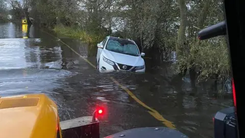 A white Nissan stuck in flood water on the edge of the road.