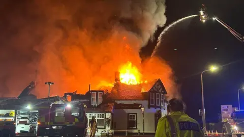 Firefighters tackle a large blaze engulfing the roof of a building at night, with flames and thick smoke rising as a fire engine’s ladder sprays water from above. A police officer and emergency responders stand in the foreground near cordons and road signs.