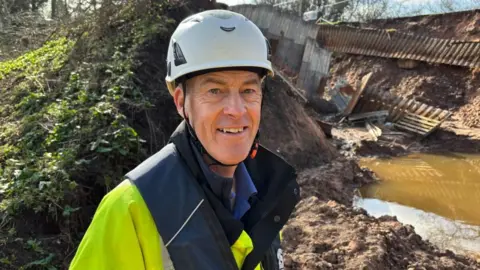 There is a man in a white hard hat, a dark blue blow-up lifejacket, and a yellow-green hi-vis jacket. He is stood behind a mound of bog-like earth, with vegetation to his left, and broken grey metal sheets to his right. There is also a dirty puddle of canal water, being held back by earth.