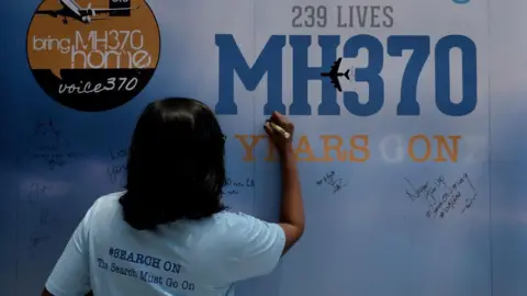 Getty Images A family member writes on a message board for passengers onboard the missing Malaysia Airlines Flight MH370 at an event in Kuala Lumpur. "The Search Must Go On," her t-shirt reads.