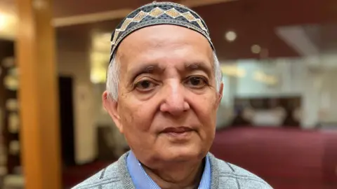 An elderly man with grey hair. wearing a hat. He is stood inside a mosque ad is looking into the camera.