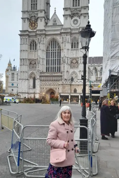 Ann Davies Ann Davies outside Westminster Abbey ahead of the carol service. She has shoulder length white hair, a pink coat, pink bag and patterned skirt. There is a railing behind her.