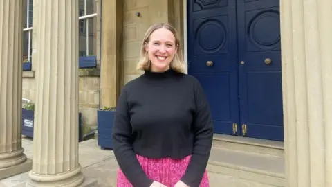 Olivia Race, a blonde woman wearing a pink skirt and black top, stood in front of a blue door with pillars.