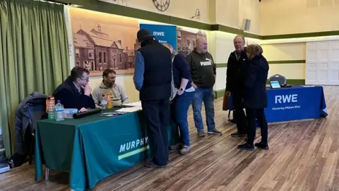 People gathered at an event inside Aylsham Town Hall. Some people are standing in front of desks, with representatives of RWE and Murphy speaking with them. The hall has archive images of the venue on the walls, with some curtains visible to the left of the image and a wood-effect floor.