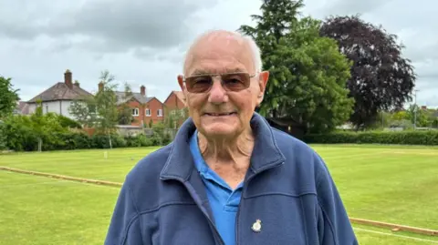 There is a man with grey hair. He has dark brown lenses on his glasses and is in his 90s. He is wearing a dark navy fleece jacket, and lighter dark blue polo shirt and a pin badge. He is stood in from of a croquet green.