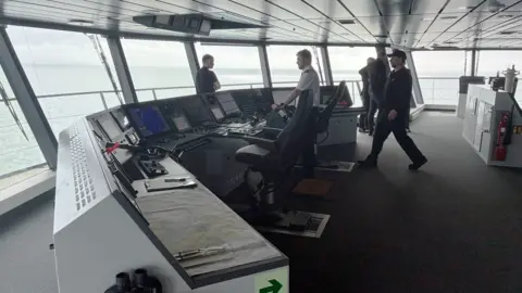 A ferry control room with several men around the room. Large windows reveal a calm still sea