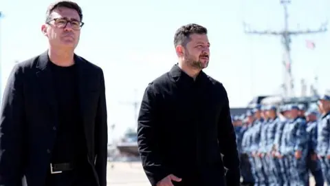 Andy Burnham and Volodymyr Zelensky walk next to each other at a naval base. Both men are dressed in black and have short, dark hair. Burnham is wearing glasses. Military personnel can be seen in the background.