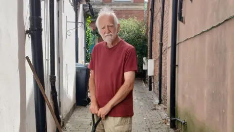 An older man in a dark red t-shirt and beige shorts. He is standing in alleyway and holding a shovel, only the handle can be seen. The man has a grey beard and short grey hair. He is pensively looking at the camera. 