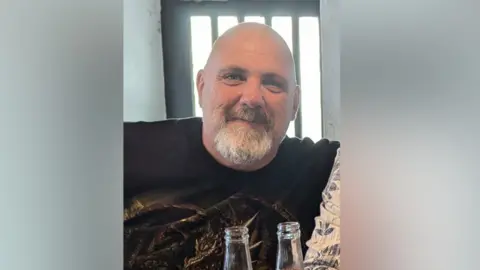 Rod Quaite Rod Quaite is bald with a grey goatee beard.He is wearing a black t-shirt and is smiling. The necks of two glass bottles can be seen at the bottom of the image - behind him is a window with bars on it.