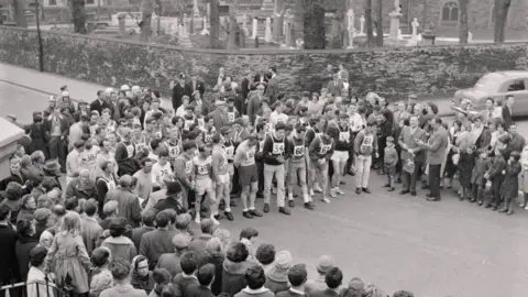  A black and white photograph of a group of people gathered around the starters of the Parish Walk. In the background is a church graveyard. 