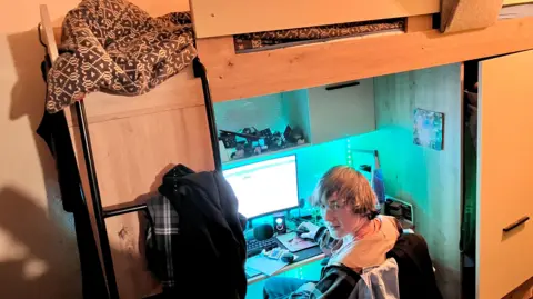 Fin sits at a desk under his bed using his computer to do his homework, he looks back toward the camera in a blue light. His desk is set up in the space under a bunk bed. A black ladder can be seen to the left of the image.