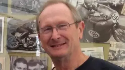 Allan Brew Allan Brew has short dark hair, is wearing glasses and a black top. He is smiling. There are black and white photos of old bikes on the wall behind him.