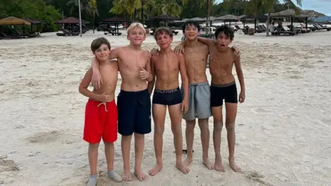 Blake Williams Five boys stand on a beach. Several of them have their arms around each other's shoulders. All are wearing swimming trunks.