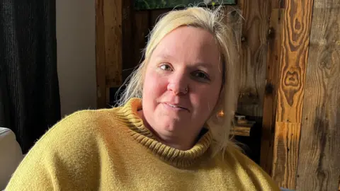 A woman with blond hair tied up in a ponytail is looking into the camera with a neutral expression. She is wearing a yellow woolen jumper and has a silver nose ring. 