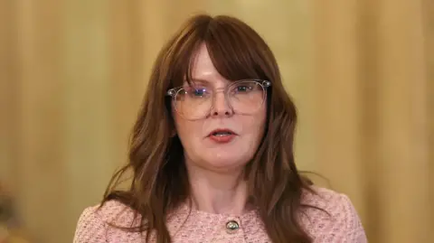 Caoimhe Archibald speaking into a microphone. She has long brown hair and is wearing a pink jacket with gold buttons and clear glasses.