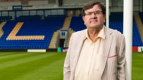 Professor Steven Broomhead, the outgoing chief executive of Warrington Borough Council, stands against the goalposts at Warrington Wolves' rugby league stadium.