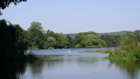 Ray Lewis The Sevenoaks Wildlife Reserve. A clear lake can be seen in the picture, surrounded by trees and shrubs.