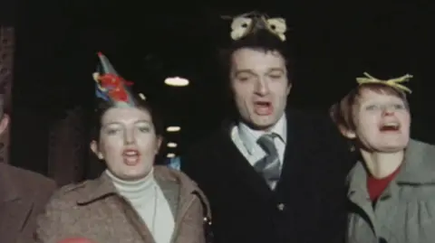 Three people, two men and a woman, wearing winter business clothes and Christmas hats, walk arm-in-arm and sing, in a picture taken in the early 1980s.