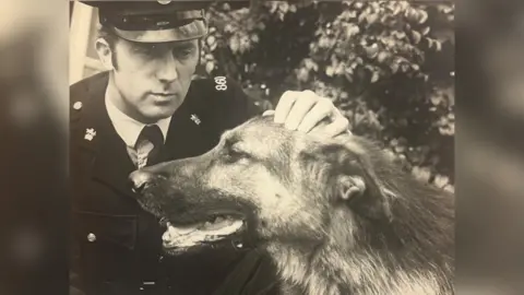 A black and white picture of PC Alan Smith with PD Shane in the 1960s. PC Smith wears an old-fashioned police uniform, with a jacket, shirt, tie and hat. PD Shane is a large German Shepherd dog. Mr Smith is captured petting Shane on the head.