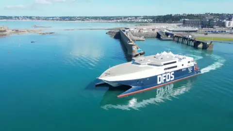 A DFDS ferry departs St Helier in Jersey on a sunny day. The water appears to be still. 