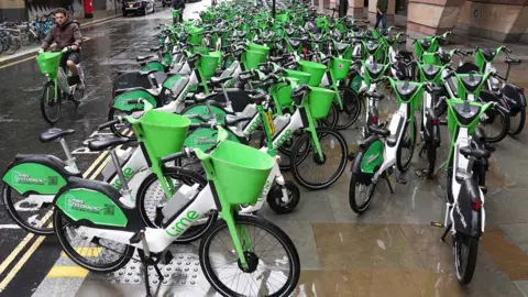 Rows of bright green Lime rental e‑bikes on a wide pavement seen extending in to the distance
