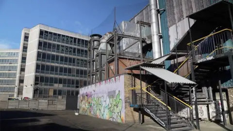 A derelict office block with broken windows and graffiti in the background with stairs up to workshops in the foreground, exposed pipework and rusting extractor fans.