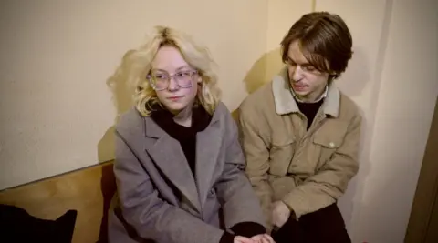 Alexander Orlov (right) and Diana Loginova (left) sit holding hands on a bench in court