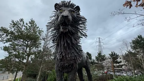 A metal sculpture of Aslan the lion in front of trees.