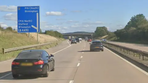 A two lane carriageway with a bridge in the distance and a large blue motorway sign on the left hand side and a number of cars on the road