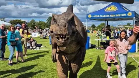 A lifelike animatronic dinosaur looks towards the camera whilst standing in a field at an outdoor event. 