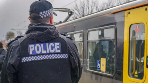 The back of a male police office who is wearing a black jacket and vest over the top with police written in white letters in a blue box across his back. There is a blue and white checked line below. He is also wearing a black cap with the same pattern. To his right there is an old Metro train which is grey with yellow doors. The sky is grey.